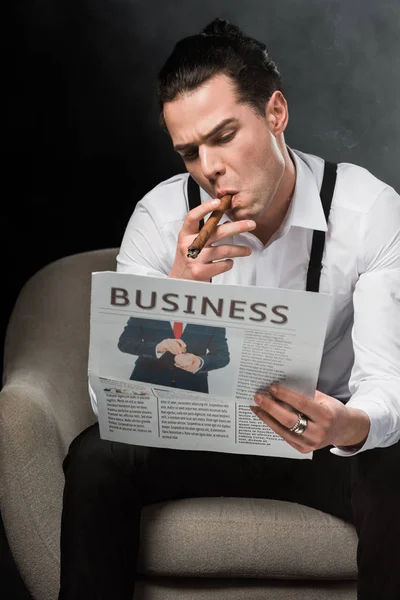Hombre serio sentado en sillón leyendo periódico de negocios y fumando cigarro en negro con humo - foto de stock