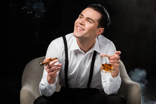 Homme heureux assis dans un fauteuil et tenant un verre de whisky et de cigare sur noir avec de la fumée — Photo de stock