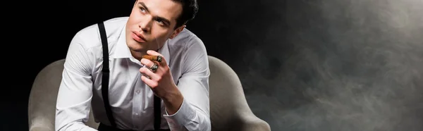 Panoramic shot of confident man holding cigar while sitting in armchair on black with smoke — Stock Photo