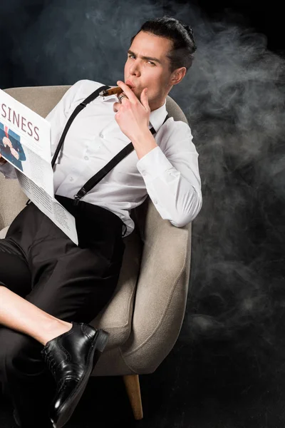 Vista aérea del hombre guapo sentado en sillón sosteniendo periódico de negocios y fumando cigarro en negro con humo - foto de stock
