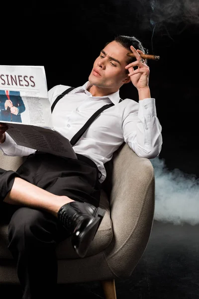 Messa a fuoco selettiva di bell'uomo seduto in poltrona leggendo giornale aziendale e tenendo sigaro sul nero con il fumo — Foto stock