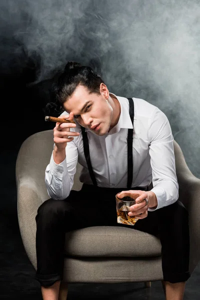 Schöner Mann blickt in die Kamera und hält ein Glas Whiskey und eine Zigarre auf schwarz mit Rauch — Stockfoto