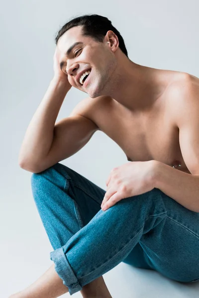 Hombre feliz y sin camisa sentado en pantalones vaqueros azules en blanco - foto de stock