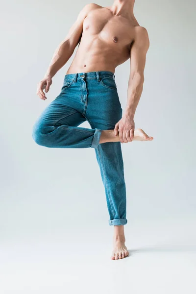 Обрізаний вид без сорочки чоловіка з дотичною ногою, стоячи в блакитних джинсах на білому — стокове фото