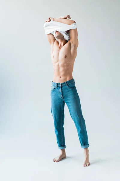 Homem muscular cobrindo rosto enquanto decolando t-shirt branca e em pé no branco — Fotografia de Stock
