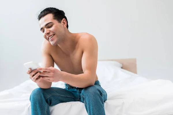 Щасливий чоловік сорочки дивлячись на смартфон, сидячи на ліжку вдома — стокове фото