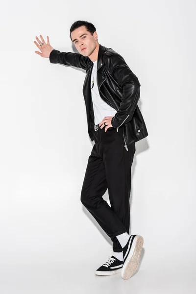 Homem bonito em jaqueta de couro preto de pé com pernas cruzadas no branco — Fotografia de Stock