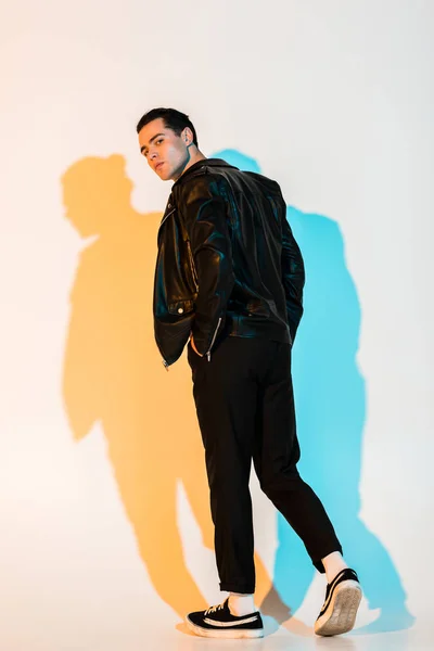 Handsome man in black leather jacket standing with hand in pocket and looking at camera on white with illumination — Stock Photo