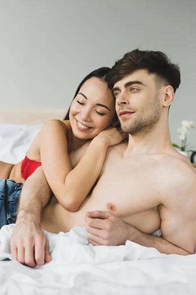 Feliz asiático mujer abrazando pensativo sin camisa novio en dormitorio - foto de stock