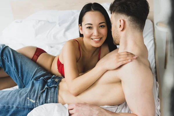 Счастливая азиатская женщина улыбается и смотрит в камеру, обнимая парня без рубашки в спальне — стоковое фото