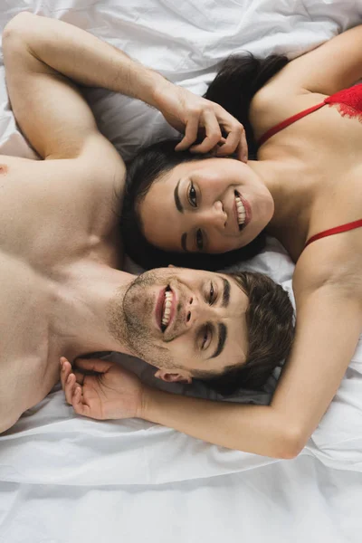 Vue de dessus du couple interracial heureux couché sur le lit, souriant et regardant la caméra — Photo de stock