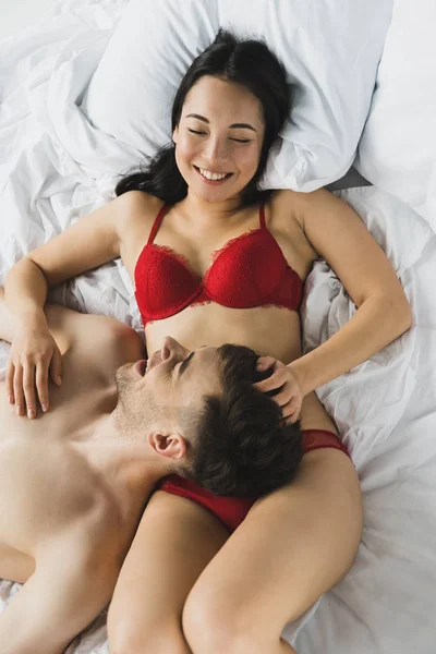 Top view of cheerful asian girl in red lingerie lying on bed near smiling boyfriend — Stock Photo