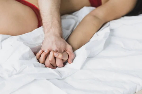 Vista recortada del hombre y la mujer tomados de la mano mientras están acostados sobre ropa de cama blanca - foto de stock