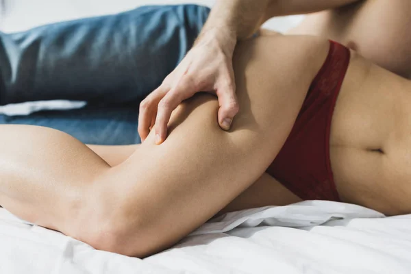 Vista recortada del hombre en pantalones vaqueros azules tocando pierna de mujer en lencería roja mientras está acostado en ropa de cama blanca - foto de stock