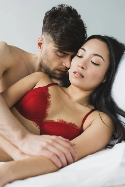 Young man kissing attractive asian girlfriend in red bra while lying on white bedding — Stock Photo