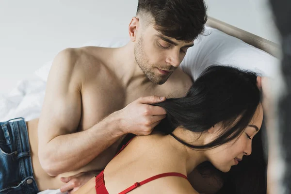 Handsome shirtless man touching hair of sexy asian girlfriend while lying on white bedding — Stock Photo