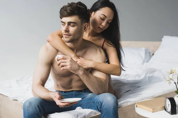 Pretty asian woman hugging handsome boyfriend sitting on bed with cup of coffee — Stock Photo