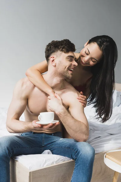 Feliz asiático mujer abrazando sonriente novio sentado en cama con taza de café - foto de stock
