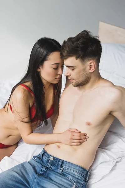 Bel homme torse nu en jeans bleus près sexy asiatique copine sur literie blanche — Photo de stock