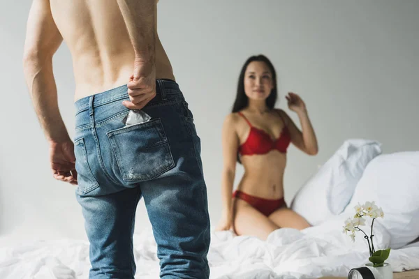 Partial view of man in blue jeans getting condom out of pocket near sexy asian girlfriend sitting on white bedding — Stock Photo