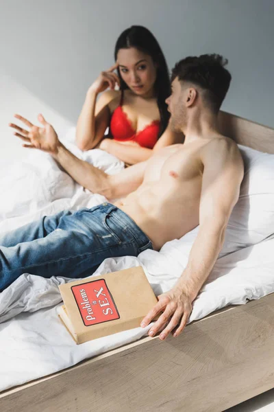 Selektiver Fokus eines hemdlosen Mannes in blauer Jeans, der Probleme beim Sex hat und mit seiner asiatischen Freundin in roter Dessous spricht, während er auf weißem Bettzeug liegt — Stockfoto
