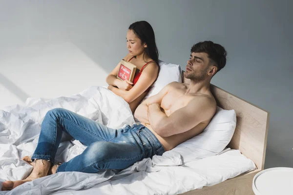 Pretty pensive asian woman holding problems in sex book while lying near offended boyfriend — Stock Photo