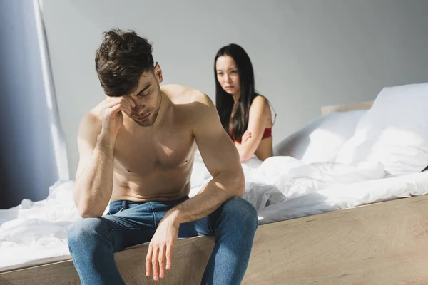 Selektiver Fokus des beleidigten, hemdslosen Mannes, der auf dem Bett in der Nähe seiner aufgebrachten asiatischen Freundin sitzt — Stockfoto