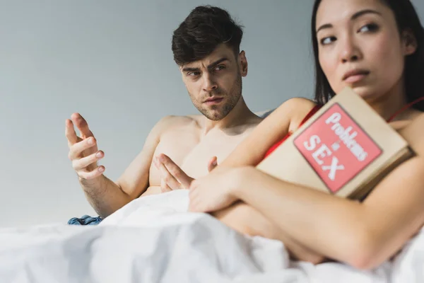 Selective focus of offended asian woman holding problems in sex book while lying near dissatisfied boyfriend — Stock Photo