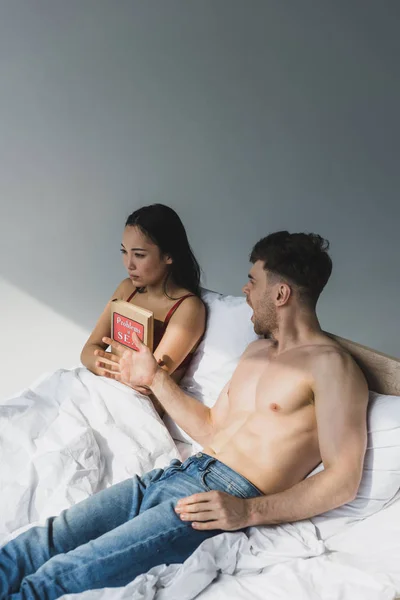 Upset asian woman holding problems in sex book while lying near quarreling boyfriend — Stock Photo