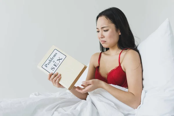Upset asian woman holding problems in sex book while sitting under white blanket — Stock Photo