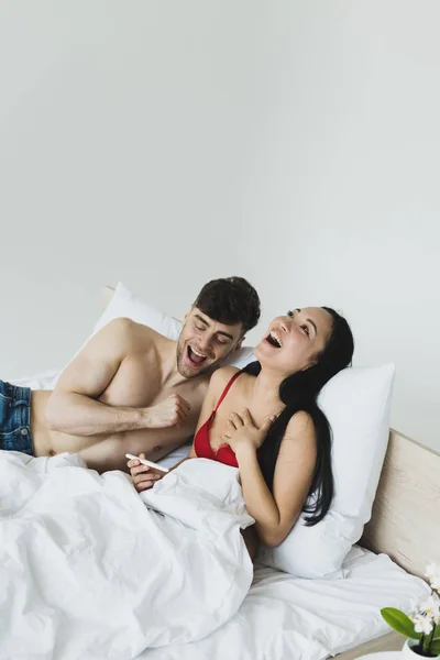 Glückliche asiatische Frau hält Schwangerschaftstest, während sie im Bett neben ihrem lächelnden Freund liegt — Stockfoto