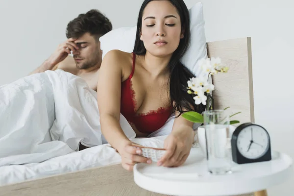 Selective focus of pretty asian woman taking birth control pills from bedside table while lying near upset boyfriend — Stock Photo