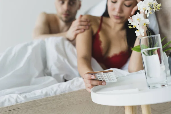 Enfoque selectivo del hombre tocando hombro de novia asiática sosteniendo píldoras anticonceptivas - foto de stock