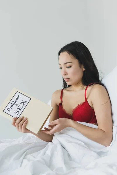 Sorprendida mujer asiática sosteniendo problemas en sexo libro mientras acostado en cama - foto de stock