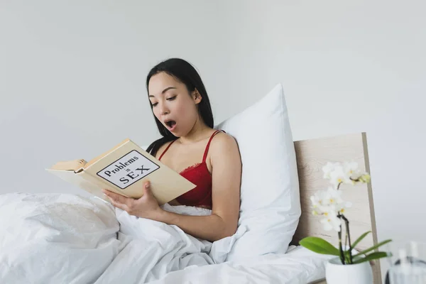 Shocked asian woman reading problems in sex book while lying in bed — Stock Photo