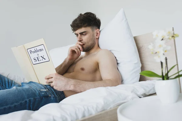 Nachdenklicher gutaussehender Mann liest Probleme in Sexbuch, während er in Blue Jeans im Bett liegt — Stockfoto