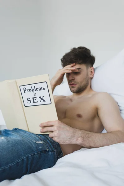 Serious shirtless man reading problems in sex book while lying in bed in blue jeans — Stock Photo