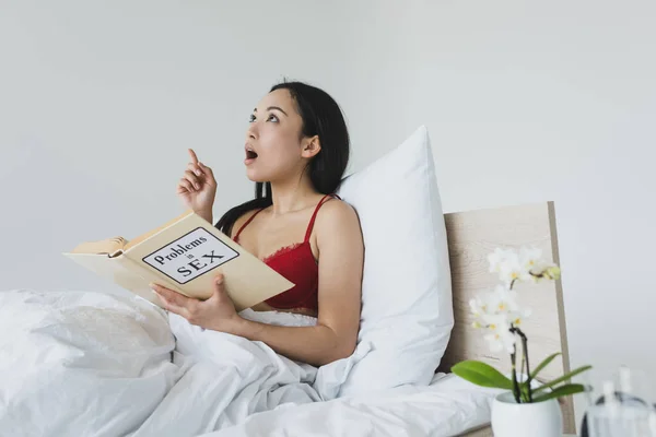 Surprised asian woman showing idea sign while reading problems in sex book — Stock Photo