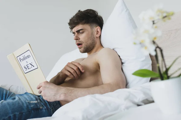 Impactado hombre guapo leer problemas en el libro de sexo mientras yacía en la ropa de cama blanca - foto de stock