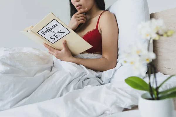 Giovane donna in lingerie rossa che legge problemi nel libro di sesso mentre giace a letto — Foto stock