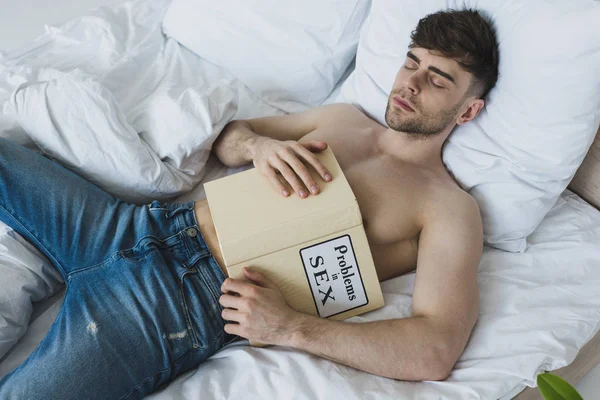 Handsome shirtless man holding problems in sex book while lying on white bedding in blue jeans — Stock Photo