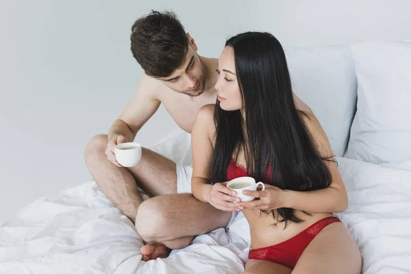 Любляча міжрасова пара сидить на білому постіллі і тримає чашки кави — стокове фото