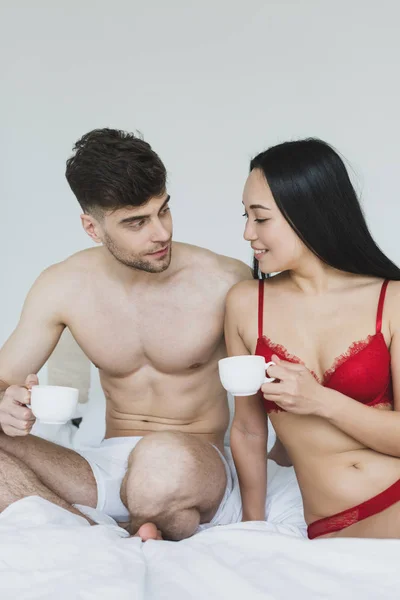 Весела міжрасова пара сидить на білому постіллі і тримає чашки кави — стокове фото