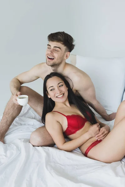 Sonriente sin camisa hombre celebración café taza mientras sentado en blanco ropa de cama cerca alegre asiático novia - foto de stock