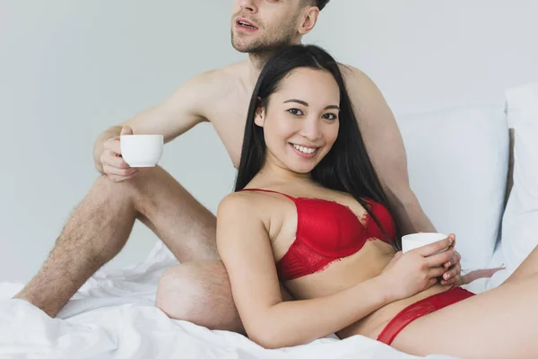 Vista parcial del hombre sin camisa sosteniendo la taza de café mientras está sentado en la ropa de cama blanca cerca de sonriente novia asiática - foto de stock