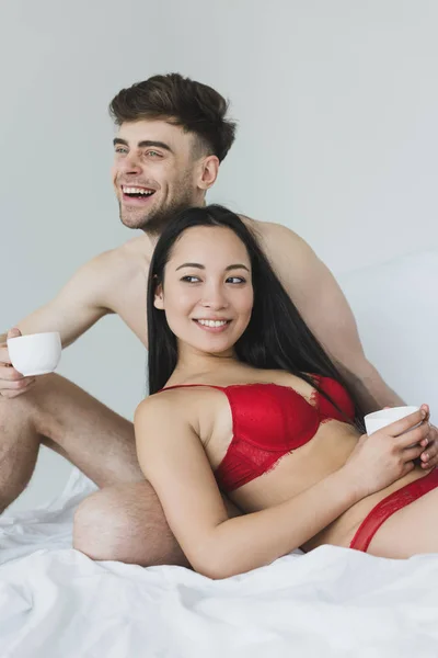 Весёлый мужчина без рубашки держит чашку кофе, сидя на белых кроватях рядом с сексуальной улыбающейся азиатской девушкой — стоковое фото