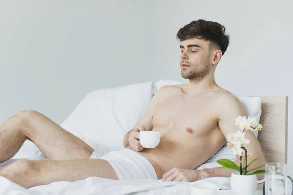 Pensive handsome man holding man holding coffee cup while lying on white bedding in underwear — Stock Photo