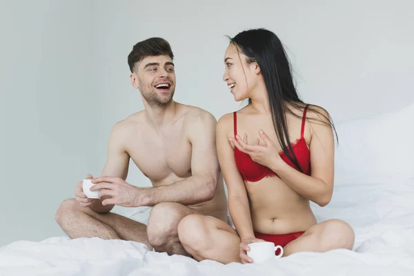 Alegre pareja interracial hablando mientras está sentado en la cama con las piernas cruzadas y la celebración de tazas de café - foto de stock