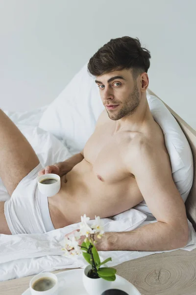 Handsome man holding man holding coffee cup and looking at camera while lying on white bedding in underwear — Stock Photo