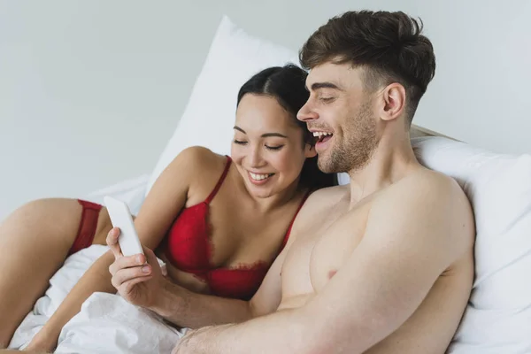 Весела міжрасова пара в нижній білизні лежить в ліжку і використовує смартфон разом — стокове фото
